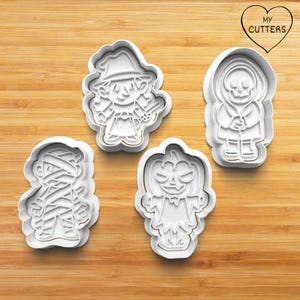 May include: Four white plastic Halloween cookie cutters, shaped like a witch, a mummy, a pumpkin, and a skeleton. The cutters are on a wooden surface.