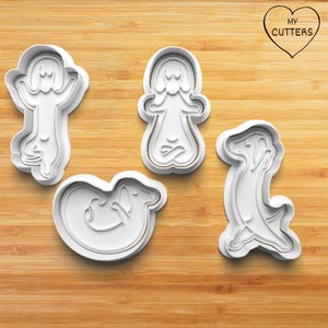 May include: Four white plastic cookie cutters shaped like dachshunds in various yoga poses. The cutters feature raised edges for cutting dough. One cutter shows a dog in a seated position, another in a standing pose, one curled, and one stretching. A heart logo with the text MY CUTTERS is in the corner.