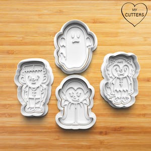 May include: Four white plastic Halloween cookie cutters, shaped like a ghost, a vampire, a werewolf, and Frankenstein's monster. The cutters are on a wooden surface.