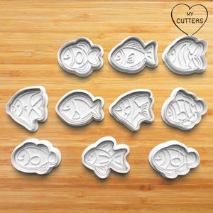 Op de afbeelding: Een set van elf witte plastic koekjesvormen in de vorm van vissen met diverse vin- en schubpatronen. Elke vorm heeft een uniek aquatisch ontwerp. Rechtsboven staat een hartvormig logo met de tekst MY CUTTERS.