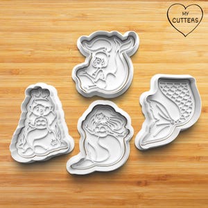 Op de afbeelding: Vier witte plastic koekjessnijders in zeemeermin vormen. De snijders omvatten een zeemeerminprinses, een zeemeermin met lang haar, een zeemeerminstaart en een zeemeermin met een baby. Op een houten oppervlak. De tekst "MY CUTTERS" staat in een hart.