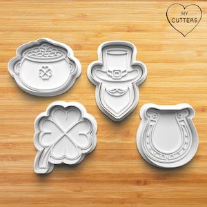 May include: Four white plastic cookie cutters shaped like a pot of gold, a leprechaun, a four-leaf clover, and a horseshoe. The cutters are on a wooden surface. The text "MY CUTTERS" is in a heart shape in the top right corner of the image.