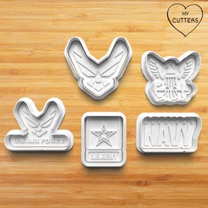 May include: Five white cookie cutters with gold trim, featuring military emblems. The cutters include the U.S. Air Force logo, an eagle, the U.S. Army star, and the words "America's Navy". The cutters are on a wooden surface.