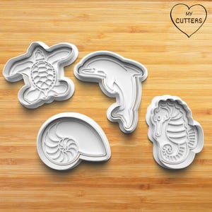 Peut inclure: Quatre emporte-pièces en plastique blanc en forme de tortue de mer, de dauphin, de coquille de nautile et d'hippocampe. Chaque moule présente un motif intérieur détaillé. Un logo en forme de cœur en haut à droite indique MY CUTTERS.