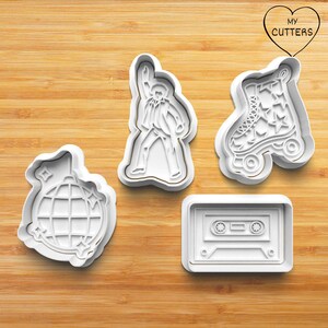 May include: Four white plastic cookie cutters shaped like a disco ball, a singer, a roller skate with stars, and a cassette tape. The cutters are on a wooden surface. The text "MY CUTTERS" is inside a heart.
