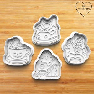 May include: A set of four white plastic cookie cutters featuring Halloween-themed amusement park designs. The shapes include a skeleton in a pumpkin teacup, a bat in a bumper car, a ghost on a roller coaster, and a mummy riding a carousel horse.