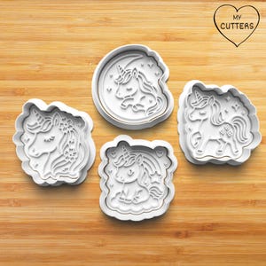 May include: Four white unicorn-shaped cookie cutters with gold trim are arranged on a wooden surface. The cutters feature different unicorn designs, including one with a crescent moon and another with a rainbow.