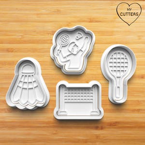 Könnte beinhalten: Satz mit vier weißen Plastik-Ausstechern in Form eines Badminton-Federballs, eines Badminton-Spielers, eines Badminton-Schlägers und eines Badminton-Netzes. Die Ausstecher befinden sich auf einer Holzoberfläche.