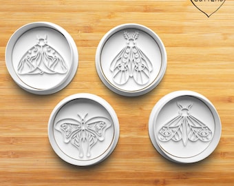 May include: A set of four white circular cookie cutters featuring intricate moth and butterfly designs with celestial patterns including moons, stars, and eyes. Each cutter has a raised edge and a detailed interior mold for shaping dough.