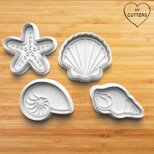May include: Four white plastic cookie cutters shaped like a starfish, a seashell, a nautilus shell, and a conch shell. The cutters are on a wooden surface.