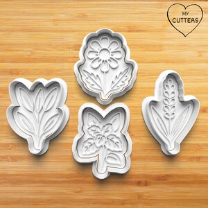 May include: Four white plastic cookie cutters in floral shapes are arranged on a wooden surface. The cutters include a daisy, a sprig of leaves, a flower with leaves, and a wheat stalk. The top right corner has a heart with the text "MY CUTTERS".