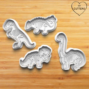 Könnte beinhalten: Vier weiße Ausstechformen aus Kunststoff in Form von Dinosaurierskeletten, darunter ein T-Rex, ein Ankylosaurus, ein Triceratops und ein Brachiosaurus. Jede Form zeigt detaillierte Skelettstrukturen zum Prägen des Teigs.