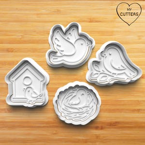 Op de afbeelding: Set van vier witte plastic koekjesvormen in de vorm van een vogelhuisje, een vogel in een nest en twee vogels op een tak. De vormen liggen op een houten oppervlak. My Cutters staat gedrukt op een hartvormig label.