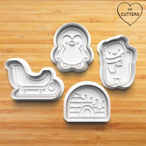 May include: A set of four white plastic cookie cutters featuring a penguin, a polar bear wearing a scarf, a sleigh, and an igloo with a brick pattern. The text MY CUTTERS is printed inside a heart shape in the upper right corner of the image.