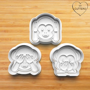 Pode incluir: Três cortadores de biscoitos de plástico branco em forma de macaco, representando as expressões não ver, não ouvir e não falar. Cada cortador possui uma borda elevada para cortar e detalhes internos para o rosto e as mãos do macaco.