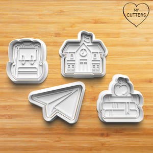 Könnte beinhalten: Ein Set aus vier weißen Kunststoff-Ausstechformen mit schulbezogenen Motiven: ein Rucksack, ein Schulgebäude mit Uhr, ein Papierflieger und ein Bücherstapel mit einem Apfel darauf. Der Text MY CUTTERS ist in einer Herzform deutlich zu erkennen.