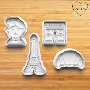 Könnte beinhalten: Vier weiße Plastik-Ausstecher in Form eines Pariser Mannes, des Eiffelturms, des Triumphbogens und eines Croissants. Die Ausstecher befinden sich auf einer Holzoberfläche.