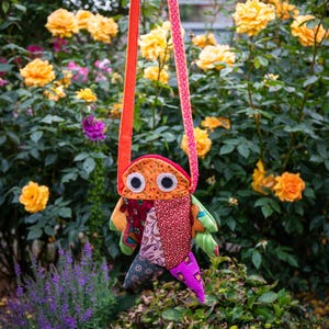 May include: A handcrafted fabric bag shaped like a stylized fish with a patchwork body featuring various patterns in red, orange, purple, and green. It has two large white circular eyes with black pupils and a long, thin, orange patterned shoulder strap.