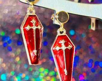 May include: A pair of coffin-shaped earrings with a red enamel finish and silver-colored trim. Each earring features a silver cross design. The earrings hang from gold-colored studs.