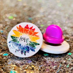 May include: A white, round pin with a rainbow sunflower design and the word "love" in black script. A bright pink pin back is attached to a white base. The background is a glittery surface.