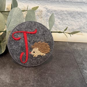 Könnte beinhalten: Ein kreisförmiger, dunkelgrauer Filzaufnäher mit einem gestickten roten Buchstaben T und einem kleinen braunen Igel. Die Stickerei zeigt ein hellbeiges Gesicht und einen dunkelbraunen, stacheligen Rücken rechts neben dem großen roten T.