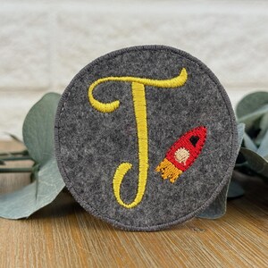 Könnte beinhalten: Ein runder, grauer Filzaufnäher mit einem gelb gestickten Buchstaben "T" und einer rot-orangenen Rakete. Der Aufnäher ist wahrscheinlich eine Applikation oder Verzierung für Kleidung oder Accessoires.