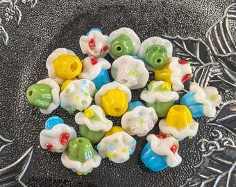May include: A collection of small, multi-colored glass beads shaped like miniature cupcakes. Each bead features a white base with a ruffled top in shades of yellow, green, blue, or white, accented with tiny colored dots or floral-like details.
