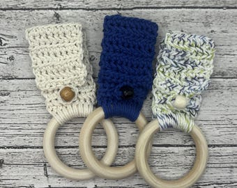 May include: Three handmade crochet towel holders with wooden rings. One is cream-colored with a wooden button, another is navy blue with a black button, and the third is a mix of blue, green, and white with a white button. The holders are on a light wood background.