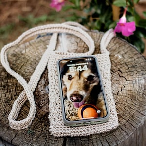 May include: A beige crocheted phone case with a matching strap rests on a tree stump. The phone displays a dog with its tongue out and the time 5:44. Pink flowers are in the background.