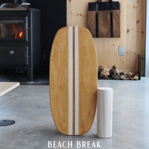 Puede incluir: Una tabla de equilibrio de madera con una franja blanca en el centro. La tabla es de color marrón claro y tiene una forma redondeada. Está colocada sobre un suelo de hormigón gris con un cilindro blanco a su lado. El texto "Beach Break" está impreso en el suelo debajo de la tabla.