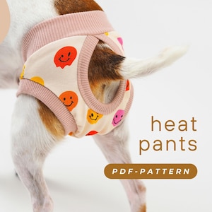 Puede incluir: Un perro blanco con pantalones de calor rosa y blanco con un patrón de caritas sonrientes. Los pantalones tienen un agujero para la cola. El texto "heat pants" y "PDF-PATTERN" es visible en la imagen.