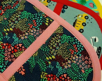 May include: A set of colourful pot holders with various patterns, including a floral design, a yellow construction truck, and a pineapple. The pot holders are made of fabric and have a pink trim.