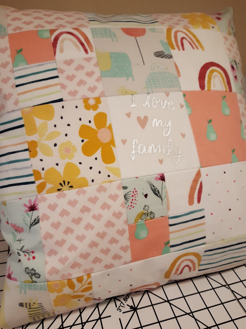 May include: A square patchwork cushion featuring various fabric patterns including rainbows, yellow flowers, pears, and hearts. A central white square displays the silver text I love my family. A ruler showing 7.6 cm is visible at the bottom edge.
