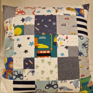 May include: A square patchwork cushion featuring a variety of fabric squares with diverse prints including construction vehicles, space themes, animals, stars, rainbows, and stripes in colours such as blue, yellow, orange, green, grey, and white.
