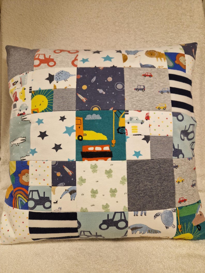 May include: A square patchwork cushion featuring a variety of fabric squares with diverse prints including construction vehicles, space themes, animals, stars, rainbows, and stripes in colours such as blue, yellow, orange, green, grey, and white.