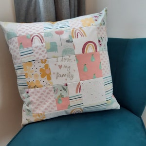 May include: A square throw cushion featuring a patchwork design of various fabric squares. The patterns include rainbows, yellow flowers, green pears, hearts, and stripes. One central square displays the text I love my family in script.