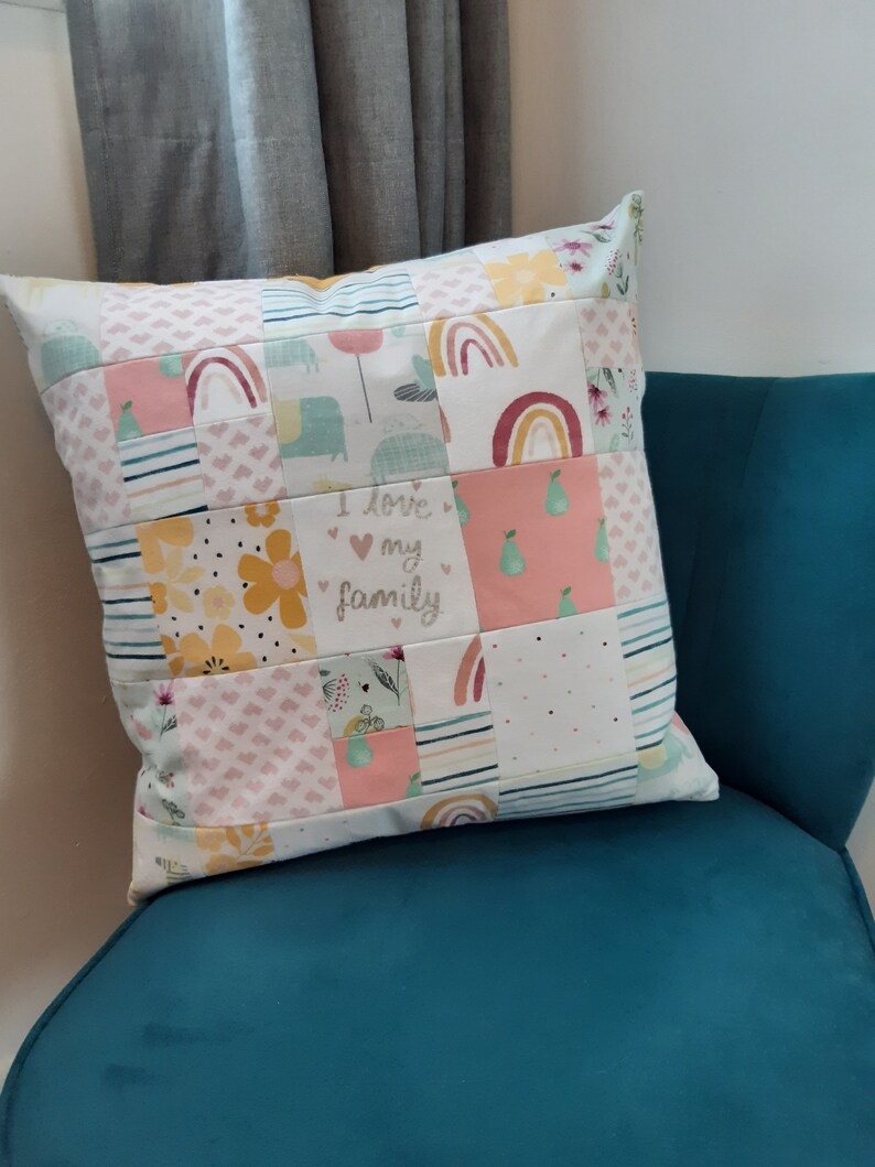 May include: A square throw cushion featuring a patchwork design of various fabric squares. The patterns include rainbows, yellow flowers, green pears, hearts, and stripes. One central square displays the text I love my family in script.