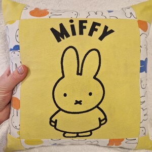 May include: A square throw cushion featuring a central yellow panel with a black outline illustration of the character Miffy and the text Miffy printed above. The border is made of white fabric with a repeating pattern of Miffy silhouettes.