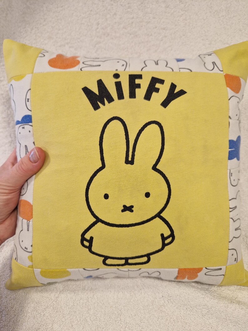 May include: A square throw cushion featuring a central yellow panel with a black outline illustration of the character Miffy and the text Miffy printed above. The border is made of white fabric with a repeating pattern of Miffy silhouettes.