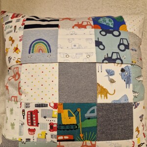May include: A square patchwork cushion constructed from various fabric squares featuring colourful prints of construction vehicles, rainbows, animals, and geometric patterns. The design includes small metal snaps integrated into the fabric seams.