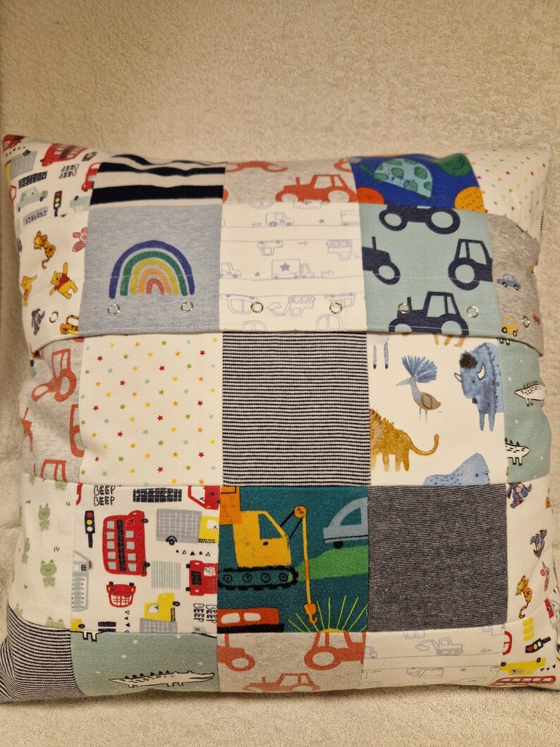 May include: A square patchwork cushion constructed from various fabric squares featuring colourful prints of construction vehicles, rainbows, animals, and geometric patterns. The design includes small metal snaps integrated into the fabric seams.