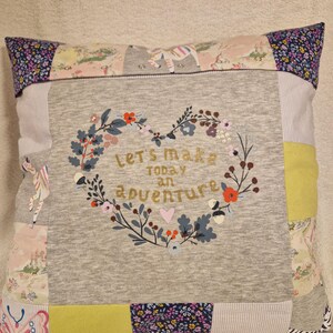 May include: A square patchwork cushion featuring a central grey panel with a heart-shaped floral wreath and the text "let's make today an adventure". The border consists of various fabric squares in patterns including butterflies, flowers, and stripes.