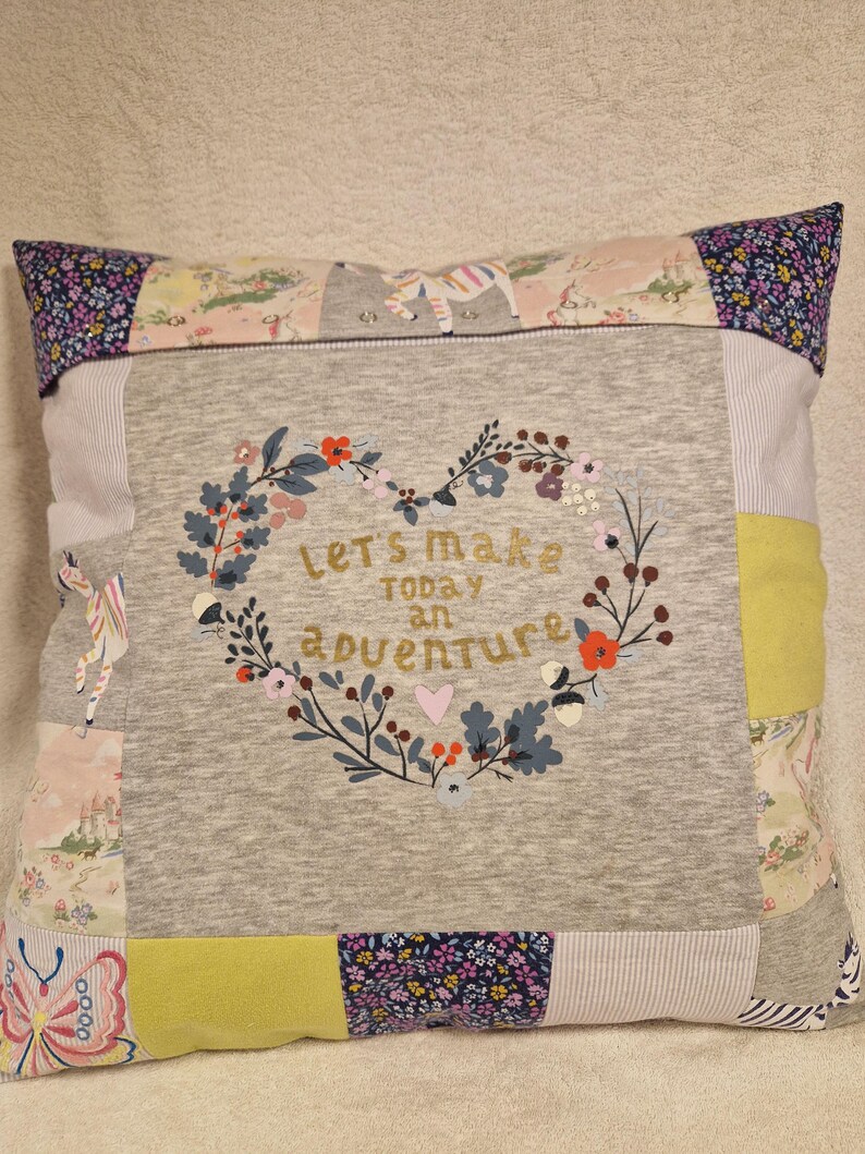 May include: A square patchwork cushion featuring a central grey panel with a heart-shaped floral wreath and the text "let's make today an adventure". The border consists of various fabric squares in patterns including butterflies, flowers, and stripes.