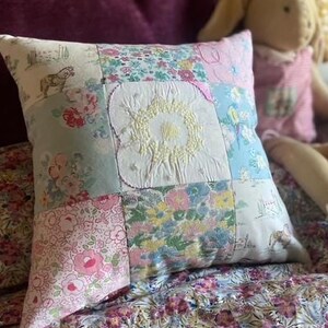 May include: A square patchwork cushion featuring various fabric squares with floral, horse, and landscape prints. The centre square is white with yellow embroidered details. The cushion is constructed from multiple textile patches sewn together.