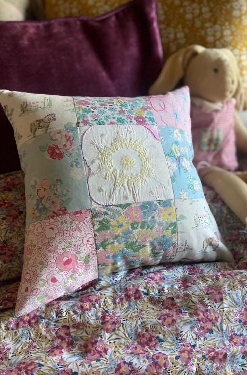 May include: A square patchwork cushion featuring various fabric squares with floral, horse, and landscape prints. The centre square is white with yellow embroidered details. The cushion is constructed from multiple textile patches sewn together.