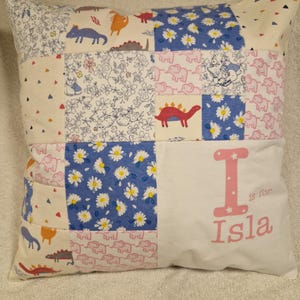 May include: A square patchwork cushion featuring a variety of fabric patterns including blue daisies, pink elephants, and colourful dinosaurs. The right side is a solid white panel with the pink text I is for Isla printed in a decorative font.