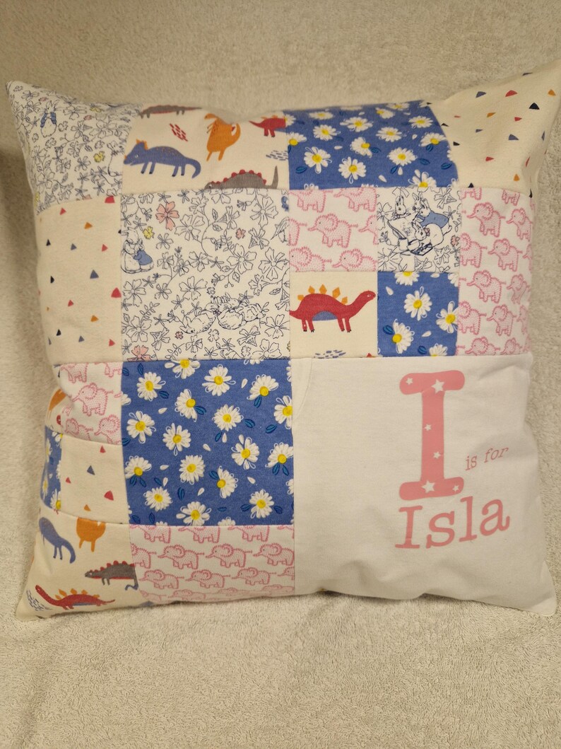 May include: A square patchwork cushion featuring a variety of fabric patterns including blue daisies, pink elephants, and colourful dinosaurs. The right side is a solid white panel with the pink text I is for Isla printed in a decorative font.