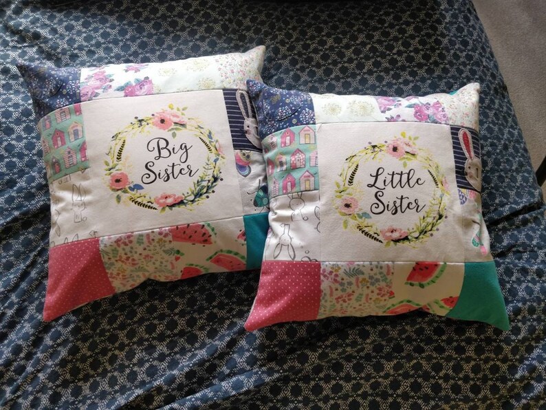 May include: Two square patchwork cushions featuring floral and fruit patterns. One cushion displays a floral wreath with the text Big Sister, and the other displays a floral wreath with the text Little Sister. The designs include rabbits, houses, and watermelons.