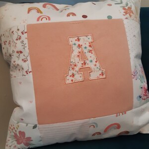 May include: A square throw cushion featuring a central solid peach-coloured fabric panel with a large, appliqued letter A in a floral print. The surrounding border consists of patchwork fabric squares decorated with rainbows, small flowers, and dots.