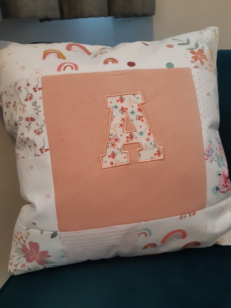 May include: A square throw cushion featuring a central solid peach-coloured fabric panel with a large, appliqued letter A in a floral print. The surrounding border consists of patchwork fabric squares decorated with rainbows, small flowers, and dots.